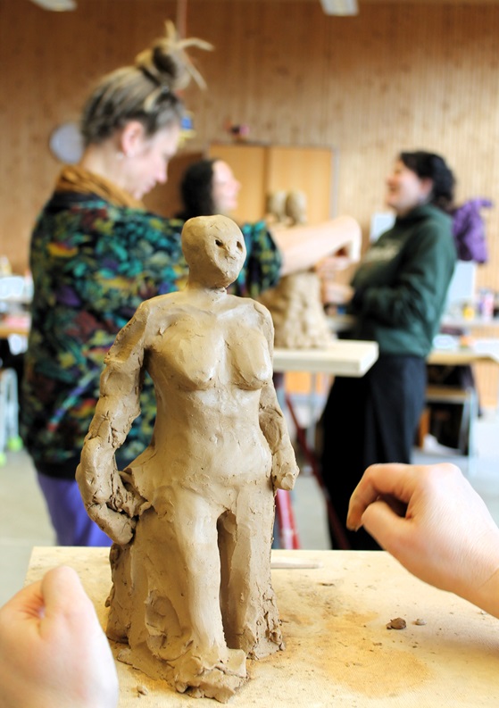 Nahaufnahme einer Tonfigur in Menschengestalt auf einem Arbeitstisch in einem Keramikkurs. Im Vordergrund sind die Hände einer Person zu sehen, die an der Skulp