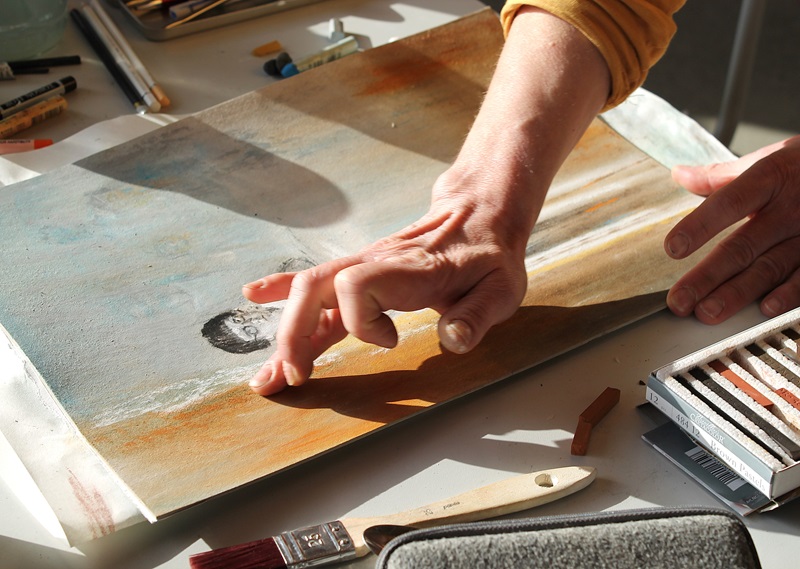 Zwei Hände verwischen mit den Fingern pastellfarbene Kreiden auf einem Blatt Papier, auf dem ein teilweise gezeichnetes Gesicht und abstrakte Farbflächen in Ora