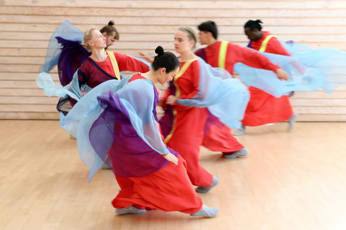 Mehrere Tänzer in bunten Gewändern führen eine dynamische Tanzbewegung in einem Raum mit Holzboden aus.
