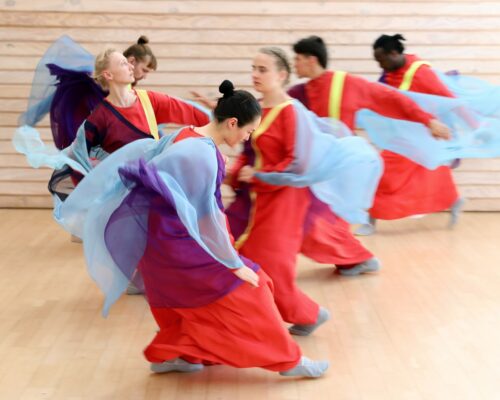 Mehrere Tänzer in bunten Gewändern führen eine dynamische Tanzbewegung in einem Raum mit Holzboden aus.