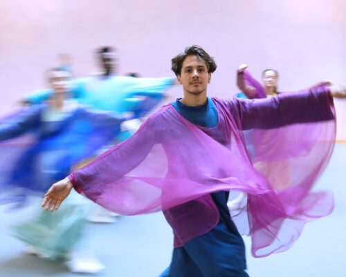 Ein Mann in einem fließenden, violetten Kleid tanzt mit ausgebreiteten Armen. Im Hintergrund sind verschwommene Gestalten in bunten Kleidern zu sehen, die ebenf