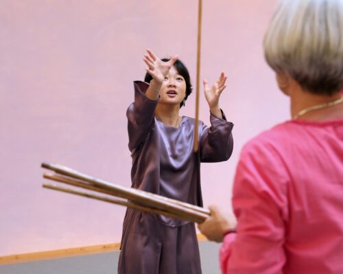 Zwei Personen in einem Raum. Eine Person trägt ein lila Seidenkleid und erhebt die Arme, während die andere Person, teilweise von hinten gezeigt, mit einem rosa