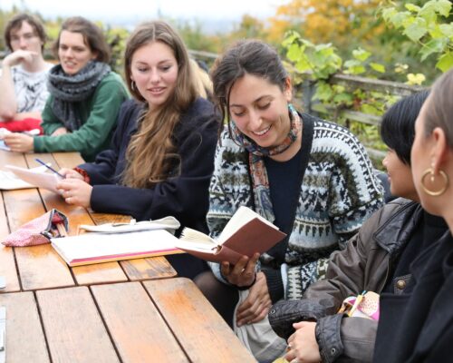 Eine Gruppe junger Menschen sitzt an einem Holztisch im Freien und diskutiert. Eine Frau in der Mitte hält ein aufgeschlagenes Buch. Alle wirken entspannt und i
