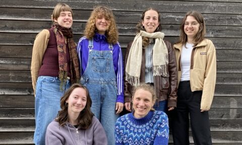 Gruppenfoto von sechs lächelnden jungen Erwachsenen vor einer Holzhauswand. Drei stehen in der hinteren Reihe, drei knien oder sitzen davor. Sie tragen lässige,