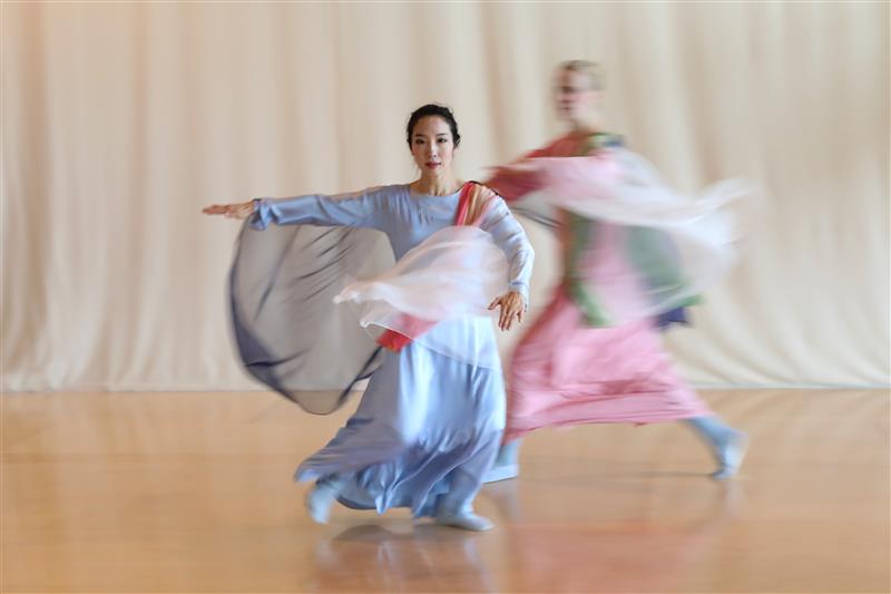 Zwei Tänzerinnen in fließenden, farbigen Gewändern bewegen sich dynamisch vor einem hellen Hintergrund. Die Bewegungen und die Kleidung erzeugen einen verschwom