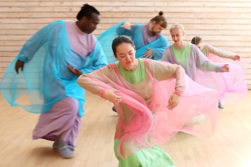 Mehrere Personen tanzen in farbenfrohen, transparenten Gewändern in einem hellen Raum mit Holzwänden. Ihre Bewegungen sind fließend.