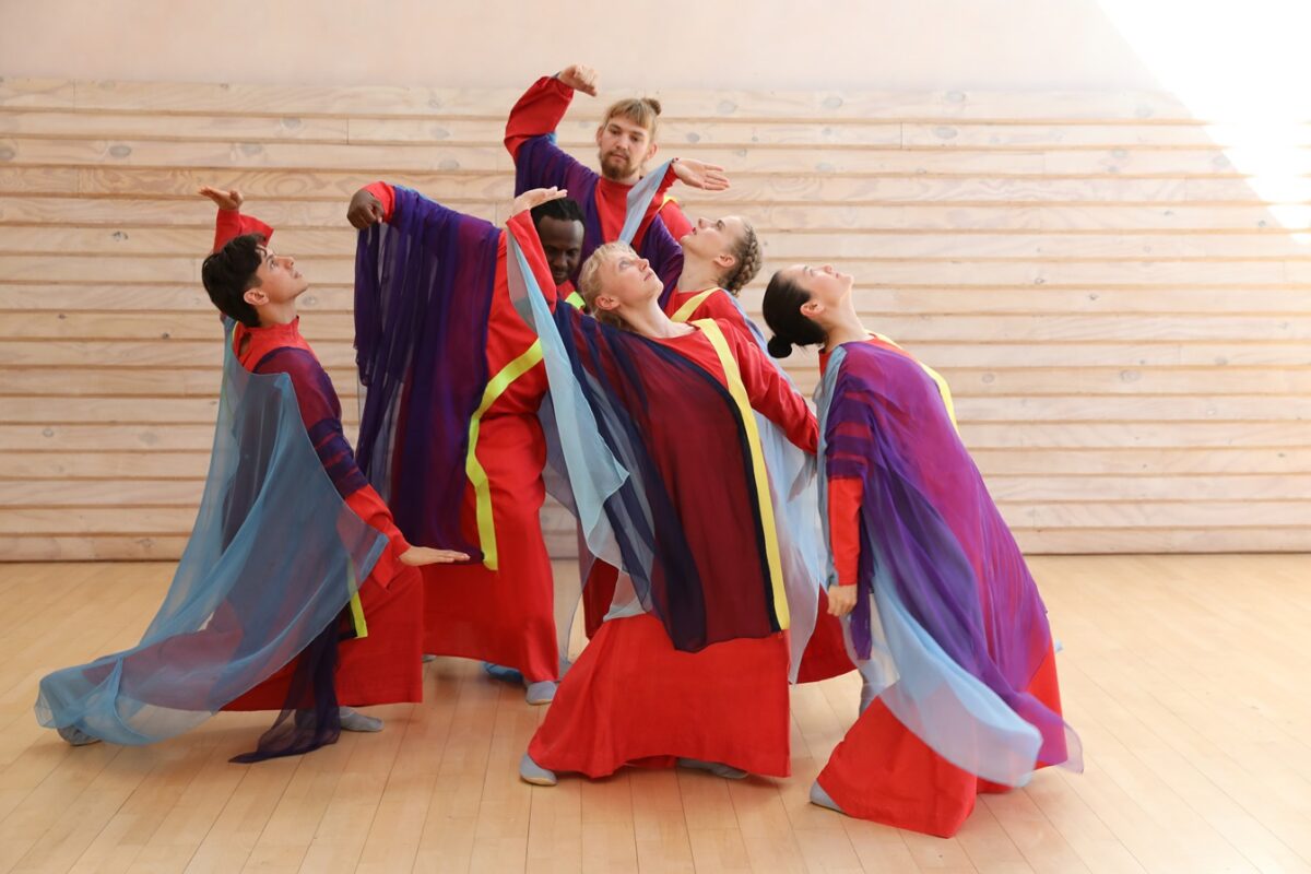 Fünf Tänzer in farbenfrohen, fließenden Kostümen führen eine Choreografie auf einem Holzboden auf.