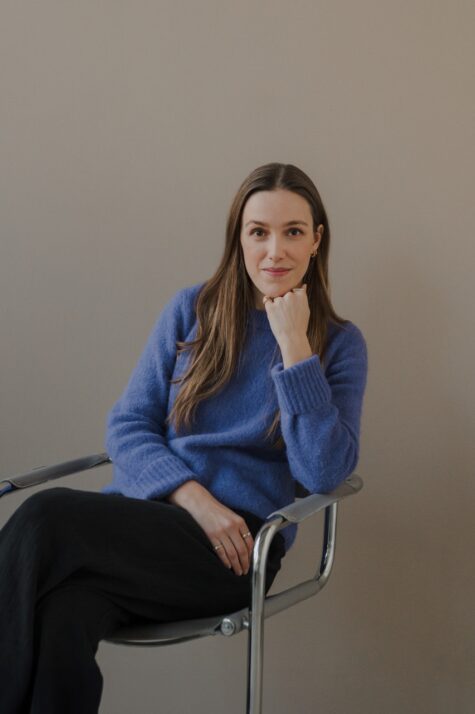 Junge Frau mit langen braunen Haaren sitzt entspannt auf einem Stuhl, trägt einen blauen Pullover und schwarze Hose und stützt ihr Kinn nachdenklich auf die Han