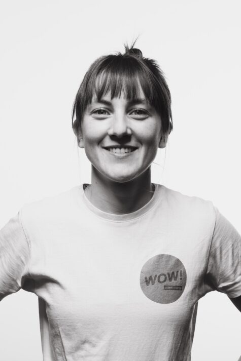Schwarz-weiß-Porträt einer lächelnden jungen Person mit zusammengebundenen Haaren und Pony, die ein T-Shirt mit dem Aufdruck „WOW! Urbane Utopien“ trägt und dir