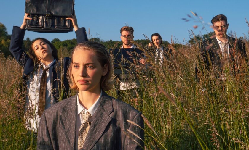 Fünf junge Erwachsene in Anzügen stehen in hohem Gras auf einer Wiese. Die Person im Vordergrund blickt ernst nach vorn, während die anderen im Hintergrund Akte