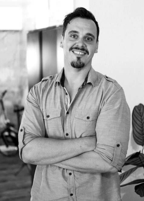 Schwarz-weiß-Porträt eines lächelnden Mannes mit kurz geschnittenem, nach oben gestyltem Haar und Kinnbart, der mit verschränkten Armen in einem modernen Innenr