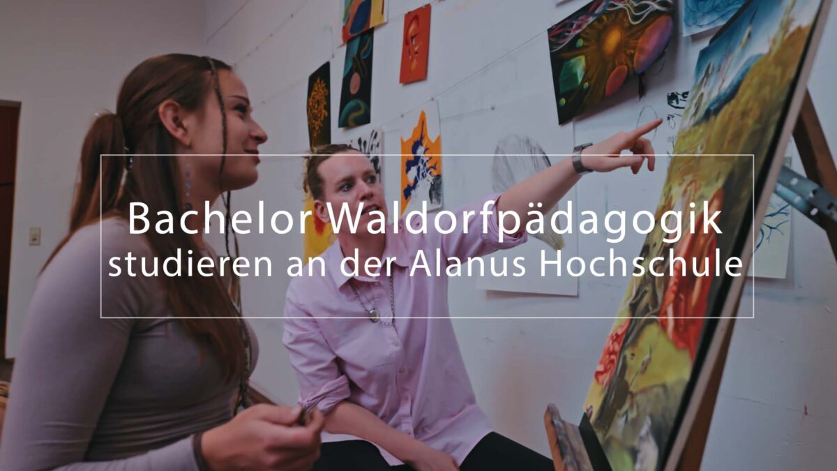 Zwei Studierende stehen in einem Kunstraum vor einer Staffelei und betrachten ein farbenfrohes Gemälde, während im Hintergrund weitere Bilder an der Wand hängen