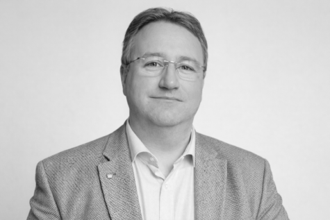 Schwarz-weiß-Porträt eines mittelalten Mannes mit Brille, kurzem Haar und leichtem Lächeln, der in einem strukturierten Sakko und Hemd vor neutralem Hintergrund