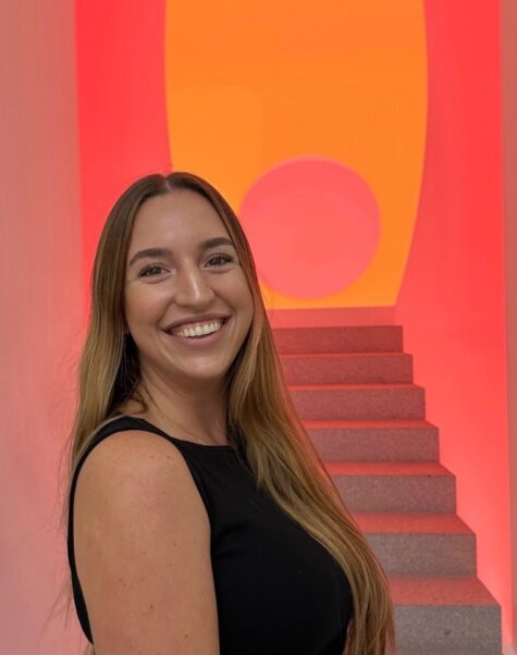 Junge Frau mit langem, braunem Haar und schwarzem ärmellosem Oberteil lächelt in die Kamera, im Hintergrund eine grau melierte Treppe vor einer leuchtenden orangen Wand.