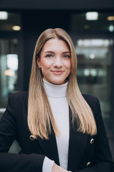 Junge Frau mit glatten, langen blonden Haaren in einem schwarzen Blazer und weißem Rollkragenpullover steht in einem modernen Bürogebäude und lächelt selbstbewu