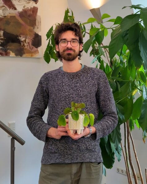 Person mit Brille und Bart steht in einem hellen Raum vor einer großen grünen Zimmerpflanze und hält einen kleinen Blumentopf mit einer runden Blattpflanze in b