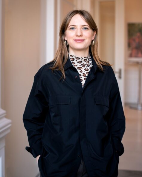 Junge Frau steht lächelnd in einem hellen Flur, trägt eine schwarze, weite Jacke über einem Rollkragenoberteil mit Leopardenmuster und goldene Creolenohrringe,