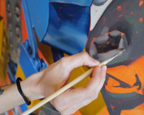 Detailarbeit mit Pinsel auf farbenfrohem Gemälde Nahaufnahme einer Hand, die mit einem feinen Pinsel Details auf einem bunten abstrakten Gemälde in Blau-, Orange- und Grautönen malt.