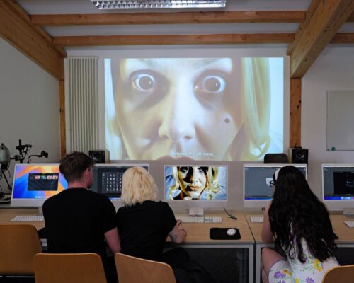 Studierende bearbeiten ein Videoprojekt im Medienlabor Drei Personen sitzen in einem Computerraum an einer Reihe von Monitoren und arbeiten, während an der Wand vor ihnen eine große Projektion eines Gesichts mit wei
