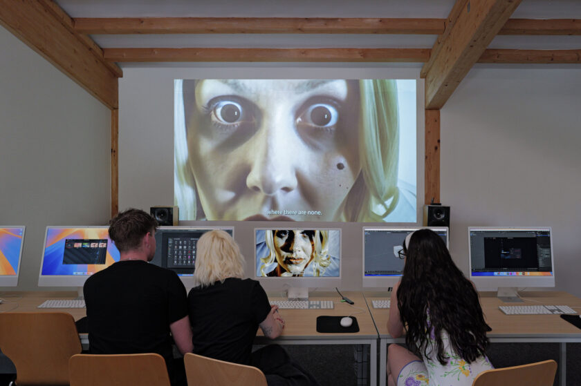 Drei Personen sitzen in einem Computerraum an einer Reihe von Monitoren und arbeiten, während an der Wand vor ihnen eine große Projektion eines Gesichts mit wei