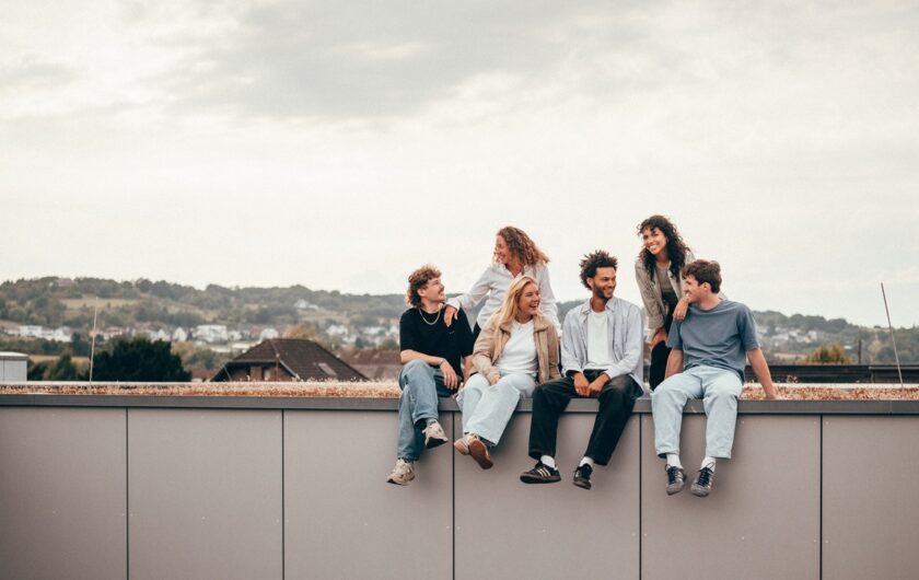 Sechs junge Erwachsene sitzen und stehen lachend auf einer Mauer mit Blick über eine Kleinstadt und hügelige Landschaft im Hintergrund.
