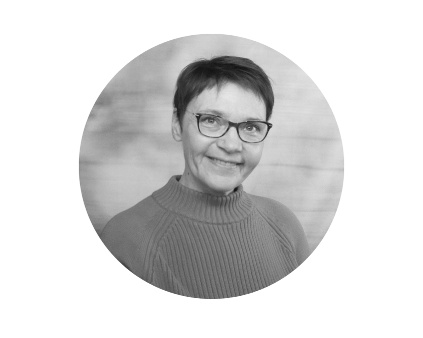 Schwarz-weiß-Porträt einer lächelnden erwachsenen Person mit kurz geschnittenen Haaren und Brille, die einen gerippten Rollkragenpullover trägt und frontal in d