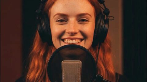 Nahaufnahme einer lächelnden Frau mit roten Haaren und großen Studiokopfhörern, die hinter einem Mikrofon in einem Tonstudio spricht oder singt.