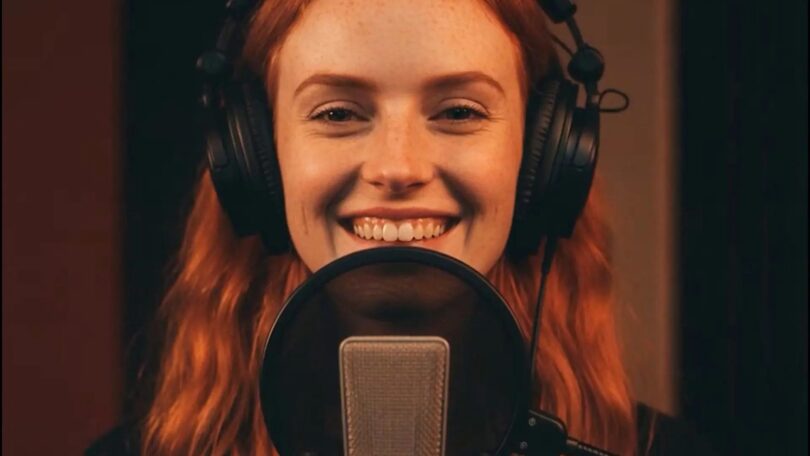 Nahaufnahme einer lächelnden Frau mit roten Haaren und großen Studiokopfhörern, die hinter einem Mikrofon in einem Tonstudio spricht oder singt.