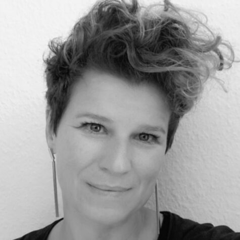Schwarz-weiß-Porträt einer lächelnden Person mit kurzem, lockigem Undercut, die seitlich in die Kamera schaut und lange, schmale Ohrringe trägt, vor einer helle