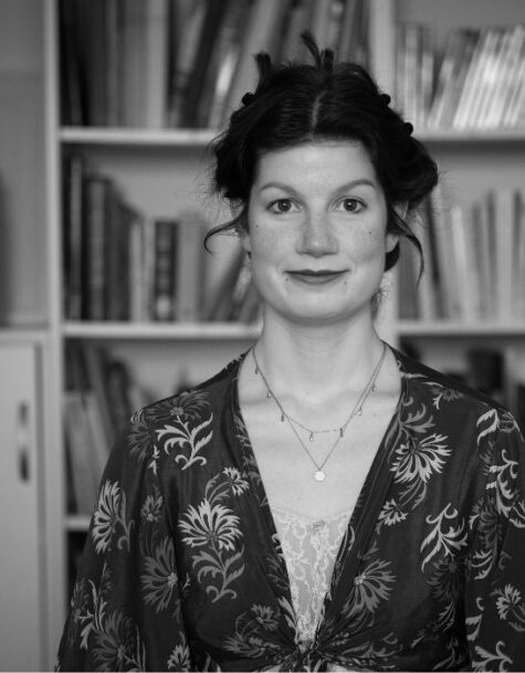 Schwarz-weiß-Porträt einer jungen Frau mit hochgesteckten Haaren, die vor einem Bücherregal steht. Sie trägt eine gemusterte Bluse mit tiefem Ausschnitt und meh