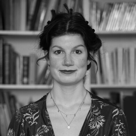 Schwarz-weiß-Porträt einer lächelnden jungen Frau mit hochgesteckten Haaren und Sommersprossen, die vor einem Bücherregal steht und in die Kamera blickt.