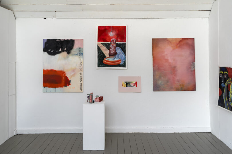 Innenansicht eines kleinen Ausstellungsraums mit weißer Wand und grauem Holzboden. Mehrere abstrakte Gemälde in kräftigen Rot-, Rosa- und Schwarztönen hängen an