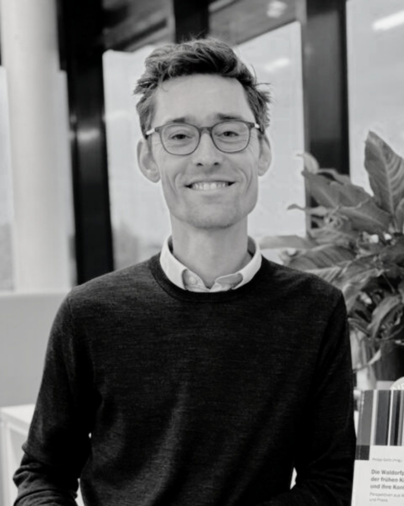 Schwarz-weiß-Foto eines lächelnden Mannes mit Brille und kurzem, leicht zerzaustem Haar, der in einem modernen Innenraum vor großen Fenstern steht. Er trägt ein