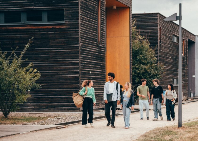 Sechs junge Erwachsene gehen lachend auf einem Weg zwischen modernen Holzgebäuden entlang. Sie tragen Freizeitkleidung und Taschen, unterhalten sich miteinander