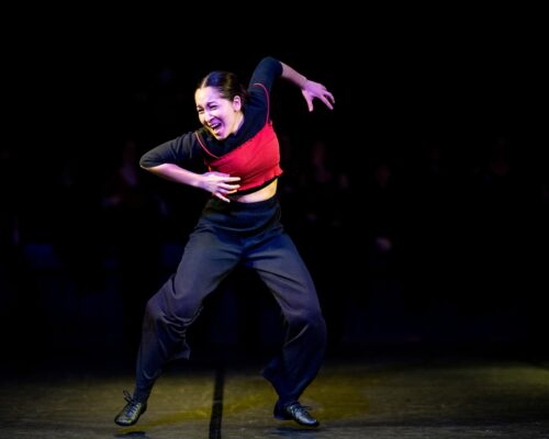 Tänzerin auf einer dunklen Bühne in dynamischer Pose, mit angespanntem Gesichtsausdruck und weit ausgebreiteten Armen, trägt ein rotes Oberteil und schwarze Hos