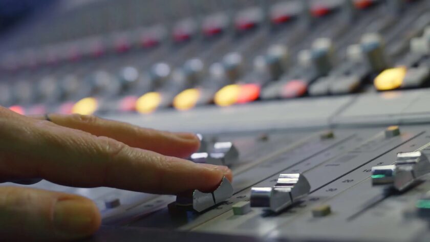 Nahaufnahme einer Hand, die an einem Mischpult einen silbernen Schieberegler bewegt, im Hintergrund leuchten unscharfe bunte Kontrolllampen.