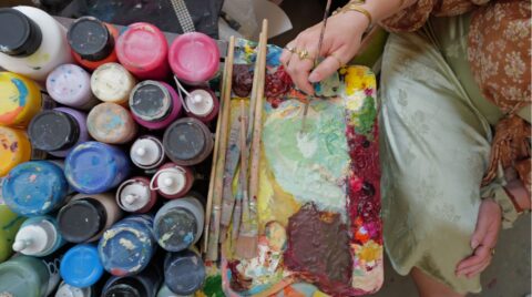 Oberansicht eines Arbeitstisches mit vielen bunten Acrylfarbflaschen, mehreren mit Farbe verschmierten Pinseln und einer voll bedeckten Mischpalette, auf der ei