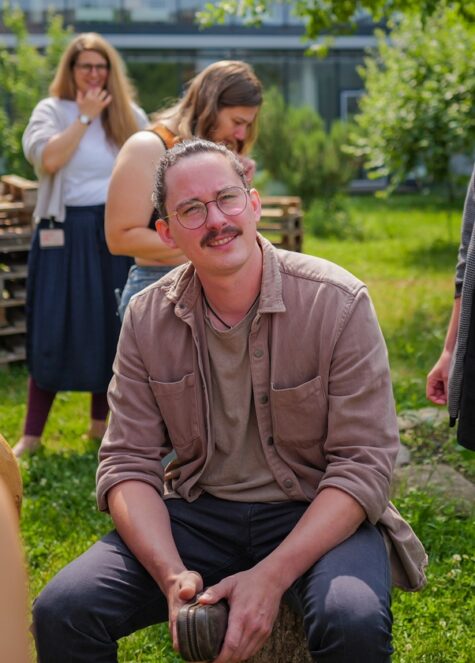 Person mit Brille und Schnurrbart sitzt entspannt auf einem Baumstumpf im Grünen und hält einen Gegenstand in den Händen, während im Hintergrund mehrere andere