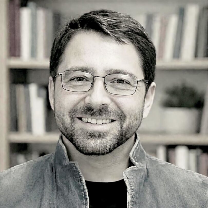 Porträt eines lächelnden Mannes mit kurzer, dunkler Frisur, Bart und Brille, der vor einem Bücherregal steht und eine offene Jeansjacke trägt.