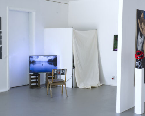 Innenraum einer modernen Kunstausstellung mit weißen Wänden. Links hängen drei kleine, schwarz‑weiße Fotografien übereinander. Davor steht ein Fernseher auf zwe