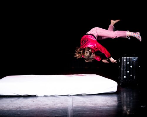 Eine Person in rosa Hose, bunten Socken und roter Jacke macht einen akrobatischen Sprung in der Luft über einer weißen Matratze auf einer dunklen Bühne; rechts