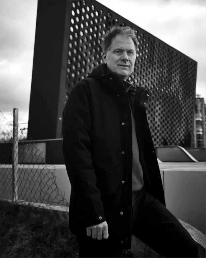 Schwarz-weiß-Fotografie eines Mannes in dunkler Jacke, der im Freien vor einer modernen Fassade mit vielen runden Öffnungen steht.