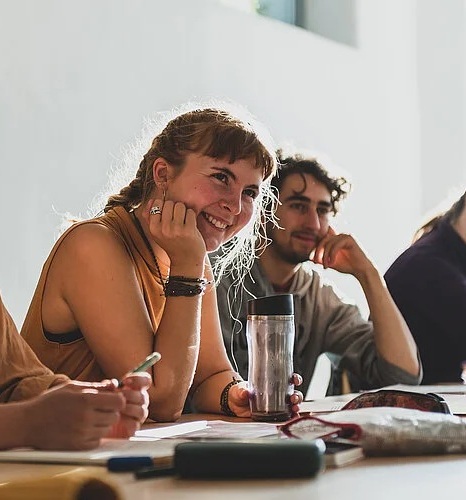 Junge Erwachsene sitzen an Tischen in einem hellen Raum, hören aufmerksam zu und lächeln, während sie Notizen machen und eine Trinkflasche auf dem Tisch steht.