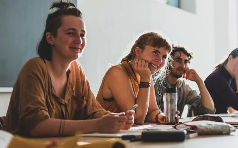 Drei junge Erwachsene sitzen lächelnd in einem hellen Seminarraum an einem Tisch, hören zu und machen sich Notizen, während vor ihnen Hefte, Stifte und eine Tri