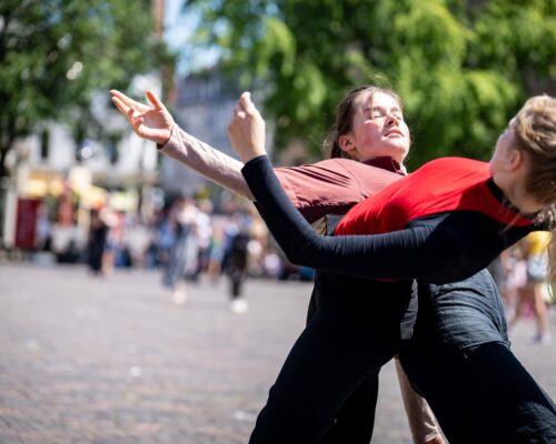 Zwei Tänzerinnen führen auf einem sonnigen Platz eine zeitgenössische Tanzperformance auf. Die eine Person in rotem Oberteil und schwarzer Hose beugt sich nach