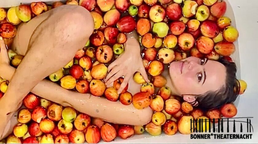 Person liegt nackt, eng zusammengerollt, in einer Badewanne voller schwimmender Äpfel und blickt lächelnd in die Kamera; unten rechts ist das Logo der Bonner Theaternacht 2026