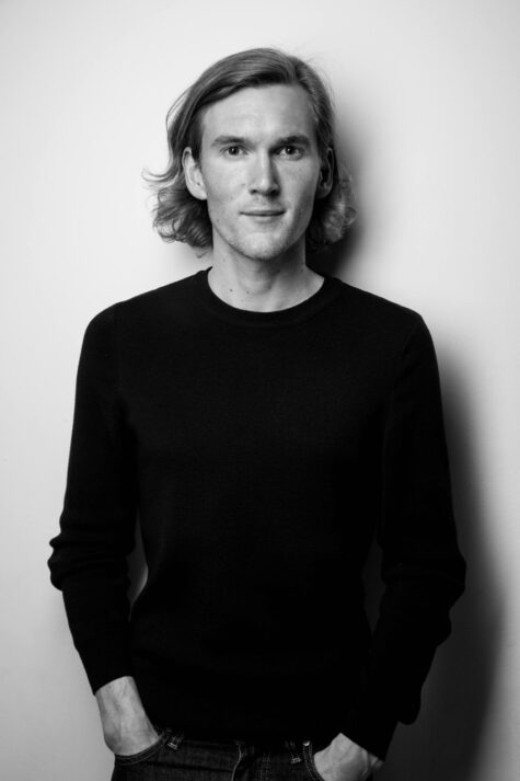 Schwarz-weiß-Porträt einer jungen erwachsenen Person mit mittellangen, glatten Haaren, die leicht lächelt und frontal in die Kamera blickt. Die Person trägt ein