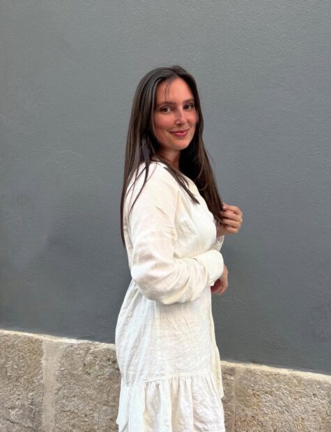 Frau mit langen braunen Haaren in einem weißen Kleid steht seitlich vor einer grauen Wand und lächelt in die Kamera.