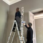 Zwischen Seidenstraße und Gegenwartskunst 3 Zwei Personen installieren ein schmales, vertikales Kunstwerk an einer hohen Wand in einem Ausstellungsraum. Eine Person steht auf einer Aluleiter und befestigt