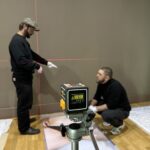 Zwischen Seidenstraße und Gegenwartskunst 5 Zwei Personen richten in einem Ausstellungsraum ein großes Foto am Boden aus. Im Vordergrund steht ein auf einem Stativ befestigter Kreuzlinien-Laser, dessen ro